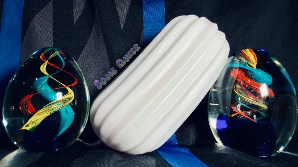 Ripple by Otouch. A white, ribbed TPE stroker. Two colorful glass eggs are on either side. The background is black, blue, and gray.